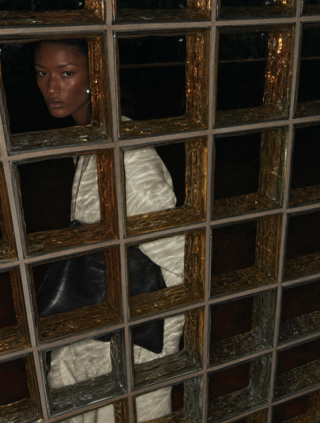 A model wearing a textured white jacket and a black shoulder bag, seen through a decorative grid of reflective gold-framed glass panels.